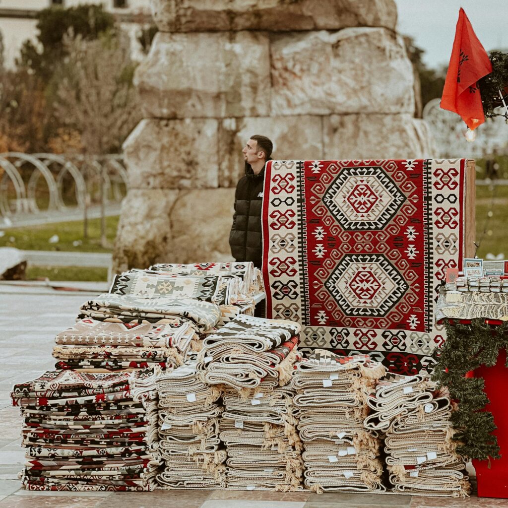 Vibrant, traditional rugs and textiles stacked at an outdoor market with a historical stone backdrop.